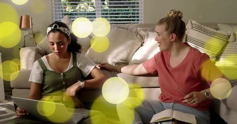 Two women sharing laptop and notebook on living room floor, studying and chatting