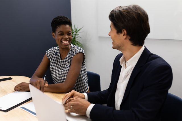 This image shows two diverse colleagues, a man and a woman, smiling at each other during a meeting in a modern office. Ideal for use in business presentations, corporate websites, and articles about teamwork, diversity, and professional collaboration.