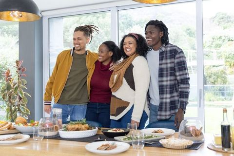 Multicultural friends standing, laughing and embracing around dining table with roast chicken