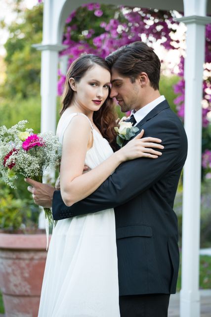 This image captures a bride and groom embracing in a beautiful garden setting, perfect for use in wedding invitations, romantic greeting cards, wedding planning websites, or bridal magazines. The lush greenery and vibrant flowers create a romantic and serene atmosphere, ideal for promoting wedding venues or floral services.