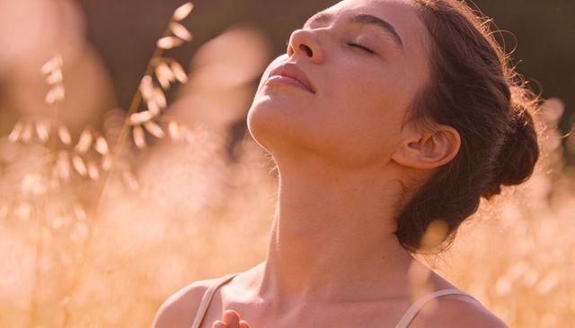 Woman soaking sunlight in golden meadow, eyes closed, experiencing mindful calm