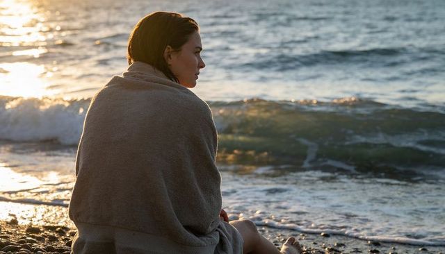 Woman wrapped in towel watching golden hour over pebbly coastline