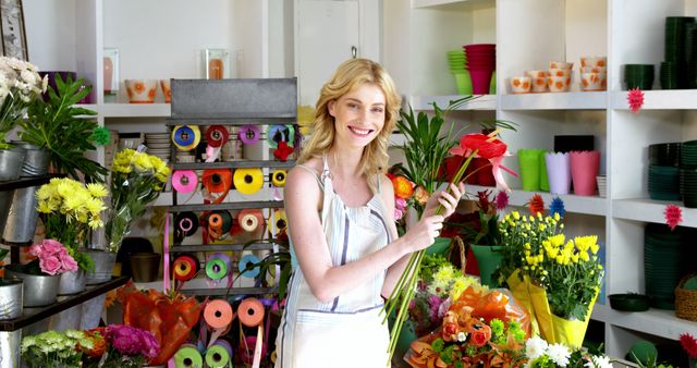 This image features a smiling florist holding flowers in a vibrant flower shop filled with various colorful blooms and supplies. Perfect for illustrating small business ownership, retail environments, or creative flower arrangements. Ideal for use in articles, blogs, or advertisements related to floristry, entrepreneurship, or retail business.