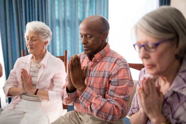 Senior man amidst women praying while sitting on chairs at retirement home