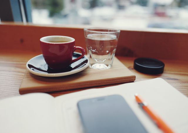 Scene shows cozy indoor café scene with red coffee cup on saucer, glass of water, open notebook, pen, smartphone resting on wooden table beside window. Suitable for themes related to mornings, work, study, writing, leisure, focus.