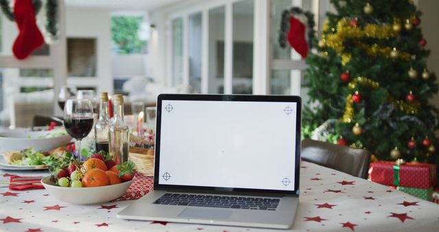 Festive Christmas scene featuring a laptop with a blank screen on table adorned with holiday decorations including fruits, wine, and a Christmas tree in the background. Suitable for holiday-related technology content, festive online greetings, or remote celebrations. Can be used in articles, blogs, or social media posts showcasing digital technology during the holidays.