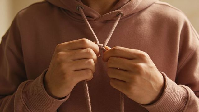 Hands tying hoodie drawstring knot on muted mauve sweatshirt, cozy detail, casual fashion