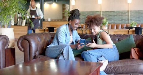 Smiling friends comparing smartphones on leather sofa in cozy modern coffee lounge