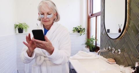 Senior woman in bathroom using smartphone with under-eye patches