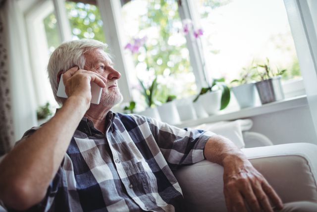 Senior man sitting on a couch, talking on a mobile phone, looking out the window. The scene is indoors with natural light coming through the window, and potted plants are visible on the windowsill. This image can be used for topics related to elderly communication, technology use among seniors, staying connected, and home life.