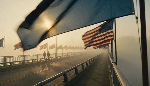 Morning fog lifting over flag-lined bridge with fluttering american flags and strolling pedestrians