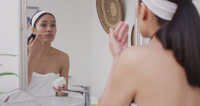 Young woman standing in front of a bathroom mirror, applying skincare cream to her face. She is wrapped in a towel and wearing a headband, suggesting a morning or evening beauty routine. This image is ideal for use in content related to personal care, skincare products, beauty tutorials, healthy skin practices, and self-care routines.