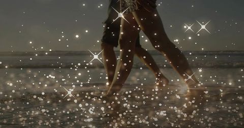Couple walking barefoot at sunset on beach shoreline with sparkling water bokeh and backlit legs