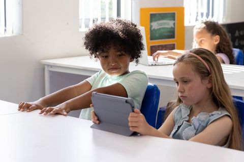 Diverse elementary students sharing tablet in bright classroom collaborating on digital learning