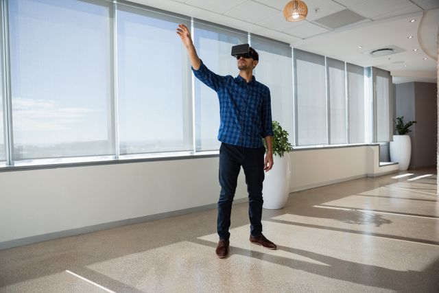 Person immersed in virtual reality in a well-lit, spacious office with floor-to-ceiling windows. Ideal for illustrating concepts related to business technology, innovation, and modern workplace environments.