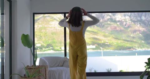 Woman standing at window in yellow overalls listening to music with ocean view