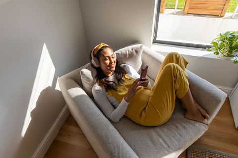 Young asian woman relaxing on gray armchair listening with headphones and using smartphone