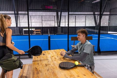 Young players chatting at wooden table next to indoor padel court with rackets