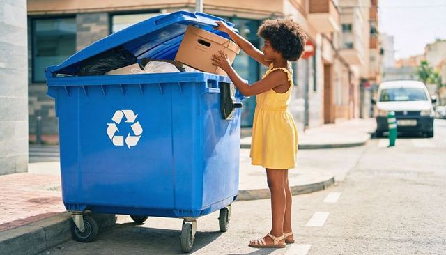 Girl in yellow dress putting cardboard into blue recycling bin on sunny urban street