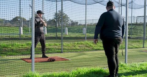 Coach coaching batter practicing swing in outdoor batting cage with netting grass glove