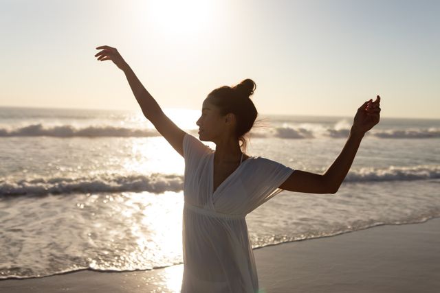 Young woman dancing joyfully on the beach during sunset, with waves crashing in the background. Perfect for use in travel brochures, wellness and lifestyle blogs, advertisements promoting beach vacations, or any content celebrating freedom and happiness.