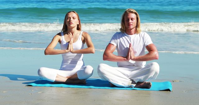 Young couple sitting cross-legged on beach near ocean practicing yoga meditation together. Both are maintaining calm and harmonious posture, wearing white outfits. Ideal for wellness, meditation, and fitness themes, promoting healthy lifestyle, relaxation, and mindfulness.