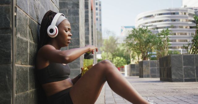 This image shows an exhausted female athlete sitting against a wall outdoors in an urban park while taking a break to drink water. She is wearing athletic clothing and headphones, making it evident she has been engaging in a physical activity, most likely running. This image is ideal for use in topics related to fitness, workout routines, hydration, urban exercising, and mental resilience during training.