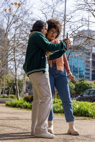 Friends checking smartphone together while walking in urban park, casual autumn fashion