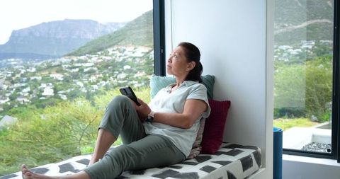 Senior asian woman reclining on window seat holding smartphone gazing over panoramic hillside view