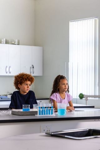 Two children sitting and watching at school lab bench with blue test tubes and goggles