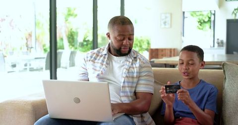 African american father teaching son online banking while sitting on couch with laptop