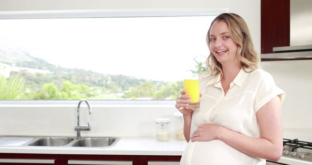 Woman in her kitchen holding a glass of fresh orange juice, smiling. Can be used for promotional material related to maternity, healthy living, morning routines, wellness tips, family life.