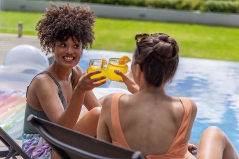 Diverse women clinking orange cocktails poolside celebrating summer relaxation in backyard
