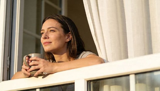 Sunlit woman leaning on windowsill holding ceramic mug, serene morning coffee moment