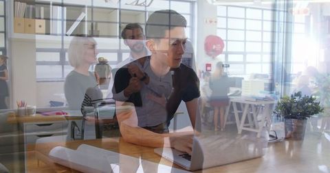 Asian Man Multitasking on Laptop Amidst Busy Office Environment
