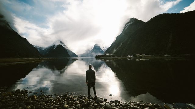 Man stands on rocky shore of calm lake, surrounded by majestic snow-capped mountains and dense clouds, creating a serene and picturesque moment. Great for travel, adventure, and nature content focusing on solitude and immersion in the natural environment.