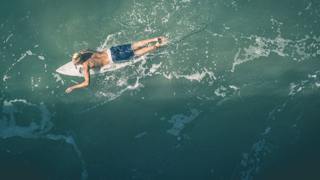A surfer paddles on a surfboard in the open ocean, creating subtle ripples on the water surface. Ideal for use in marketing materials related to surfing, adventure sports, beach vacations, and outdoor activities. Perfect for websites or blogs focused on surfing, active lifestyles, or summer recreation.