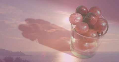 Double exposure glass of red grapes over sunlit coastal sunset creating dreamy reflection