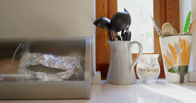 Kitchen utensils sit in a ceramic holder by a window, with copy space. Sunlight streams in, highlighting the cozy, domestic setting of a well-used kitchen space.