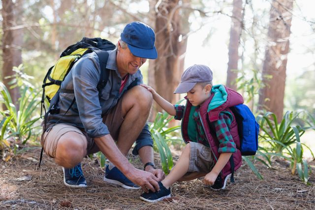 Mature father tying shoelace for young son during a hike in a forest. Both wearing casual outdoor clothing and backpacks. Ideal for use in parenting articles, outdoor activity promotions, family adventure advertisements, and travel blogs focusing on family bonding and nature exploration.