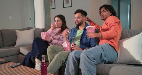 Diverse friends enjoying cozy movie night on gray sofa with popcorn and bottled drinks