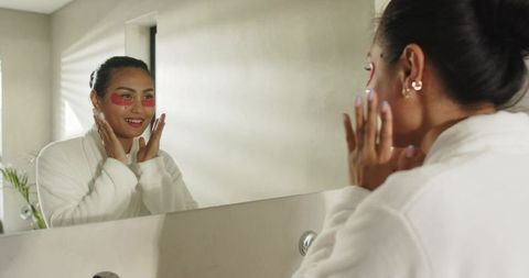 Asian woman wearing robe smiling and adjusting red under-eye patches at bathroom mirror