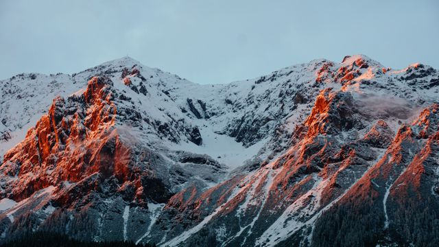 Snow-covered mountain peaks illuminated by the orange glow of sunrise, capturing the dramatic beauty of nature. Ideal for use in nature articles, travel advertisements, outdoor adventure blogs, and landscape photography galleries.