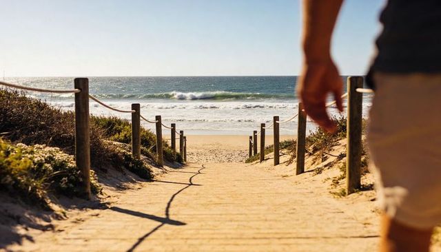 Sunlit dune pathway leading to ocean, man walking in shorts at right frame, coastline escape
