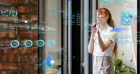 Businesswoman analyzing holographic data dashboards on glass partition in modern brick office