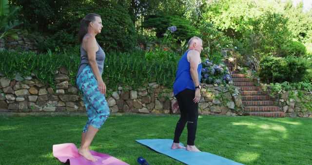 Two women practicing yoga outdoors on mats in a lush garden with stone steps in the background. Ideal for promoting outdoor fitness, wellness activities, and nature retreats.