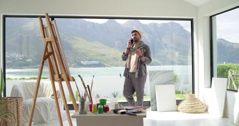 Indian artist talking on phone in bright waterfront studio with easel and paints
