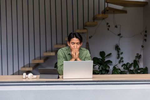 Asian man working from home on laptop and tablet looking stressed in minimalist workspace