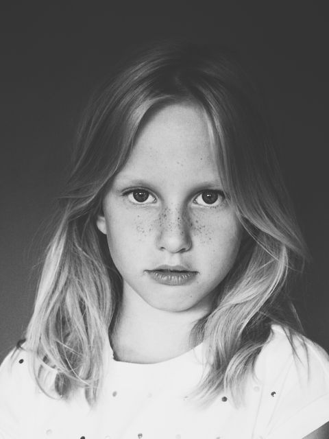 A serious young girl gazes directly at the camera, creating an introspective and somber feel. This black and white portrait highlights her fine freckles and blonde hair, capturing a timeless essence. Ideal for use in publications related to childhood, emotions, personal stories, and more.