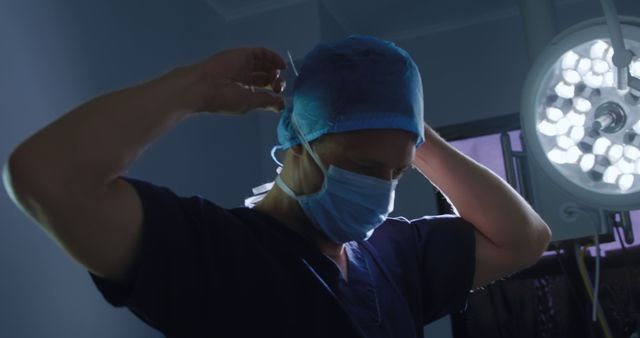 Surgeon adjusting surgical cap and mask under bright surgical lights in operating room provides an authentic glimpse into a medical professional preparing for surgery. Useful for healthcare websites, medical blogs, educational material, or advertisements promoting hospital services and medical tools.